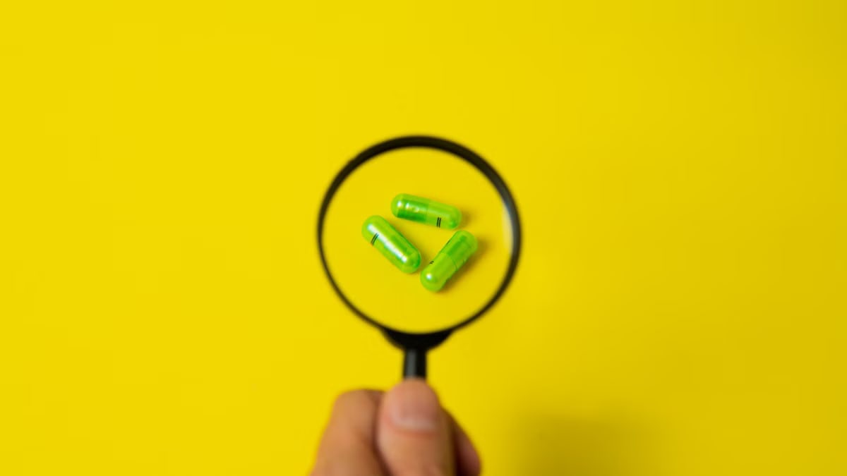 Top view of crop unrecognizable person holding magnifier over green pills placed on bright yellow surface in studio
