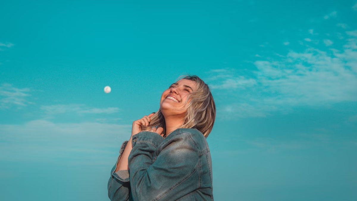 A happy woman in a denim jacket smiles under a vibrant blue sky with a visible moon.